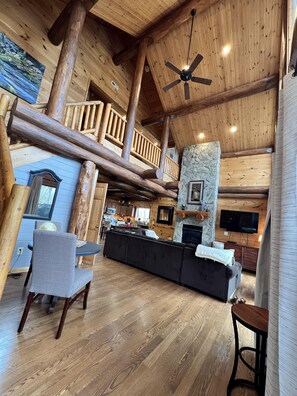 A spacious log cabin interior with a large living area, a stone fireplace, a ceiling fan, and a television mounted on the wall.
