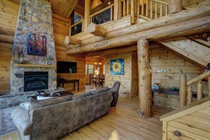 A log cabin interior with a stone fireplace, a large sofa, a dining area, and a staircase.