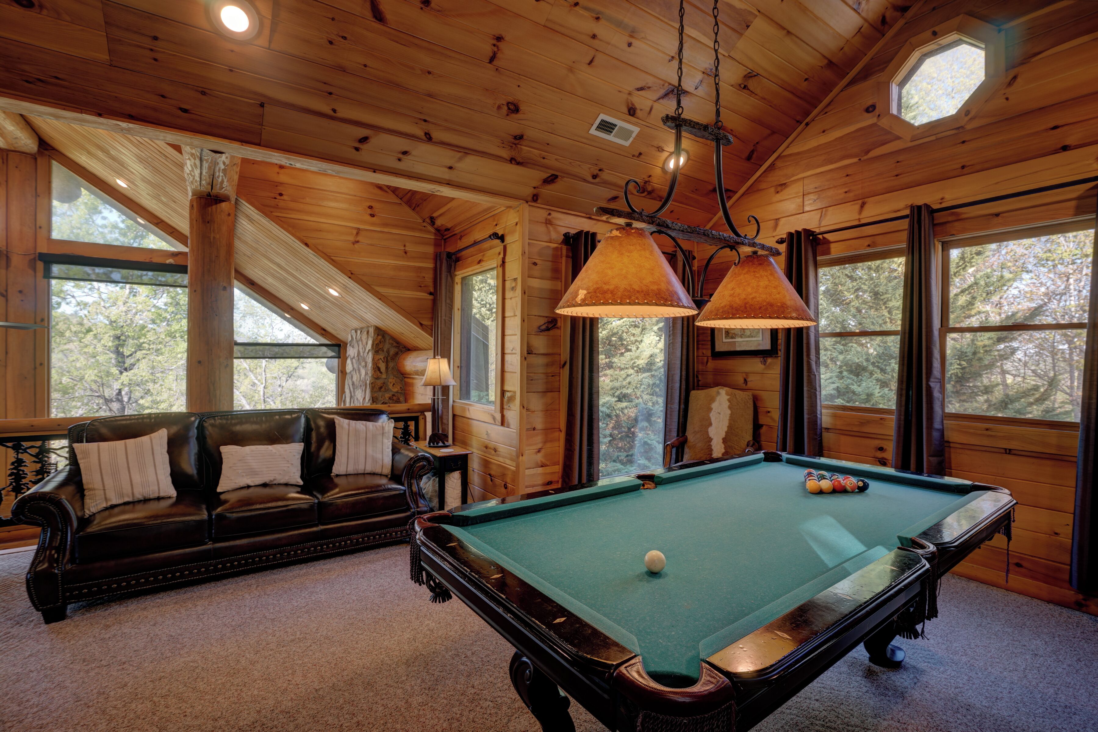 A wooden interior with a pool table, leather sofa, and large windows.
