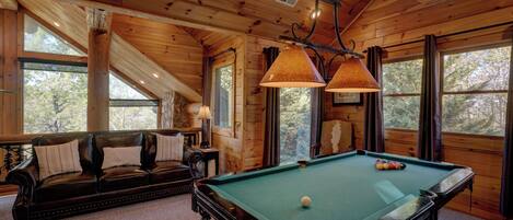 A wooden interior with a pool table, leather sofa, and large windows.