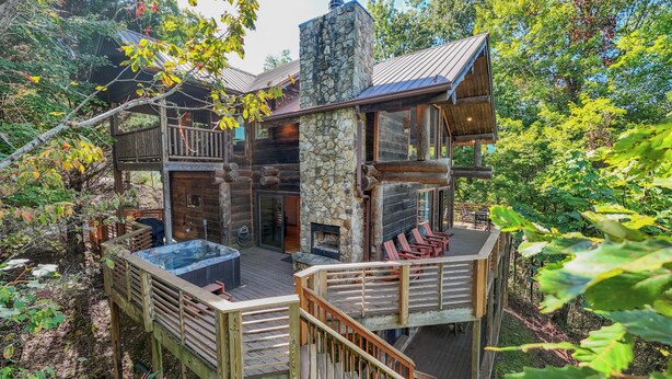 Une cabane en rondins avec une cheminée en pierre, une terrasse en bois et un espace extérieur aménagé avec des sièges, le tout entouré d’arbres.