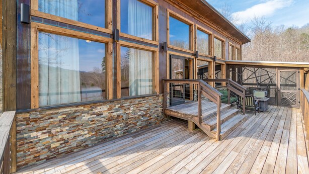 Una terraza de madera con una escalera que conduce a un espacio cubierto, ventanas amplias y un muro de piedra.