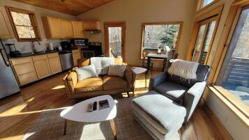A cozy living area with a sofa, armchairs, and a coffee table. A kitchen with wooden cabinets and appliances is visible in the background.