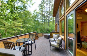 Una terraza de madera con mobiliario exterior, rodeada de un bosque frondoso.