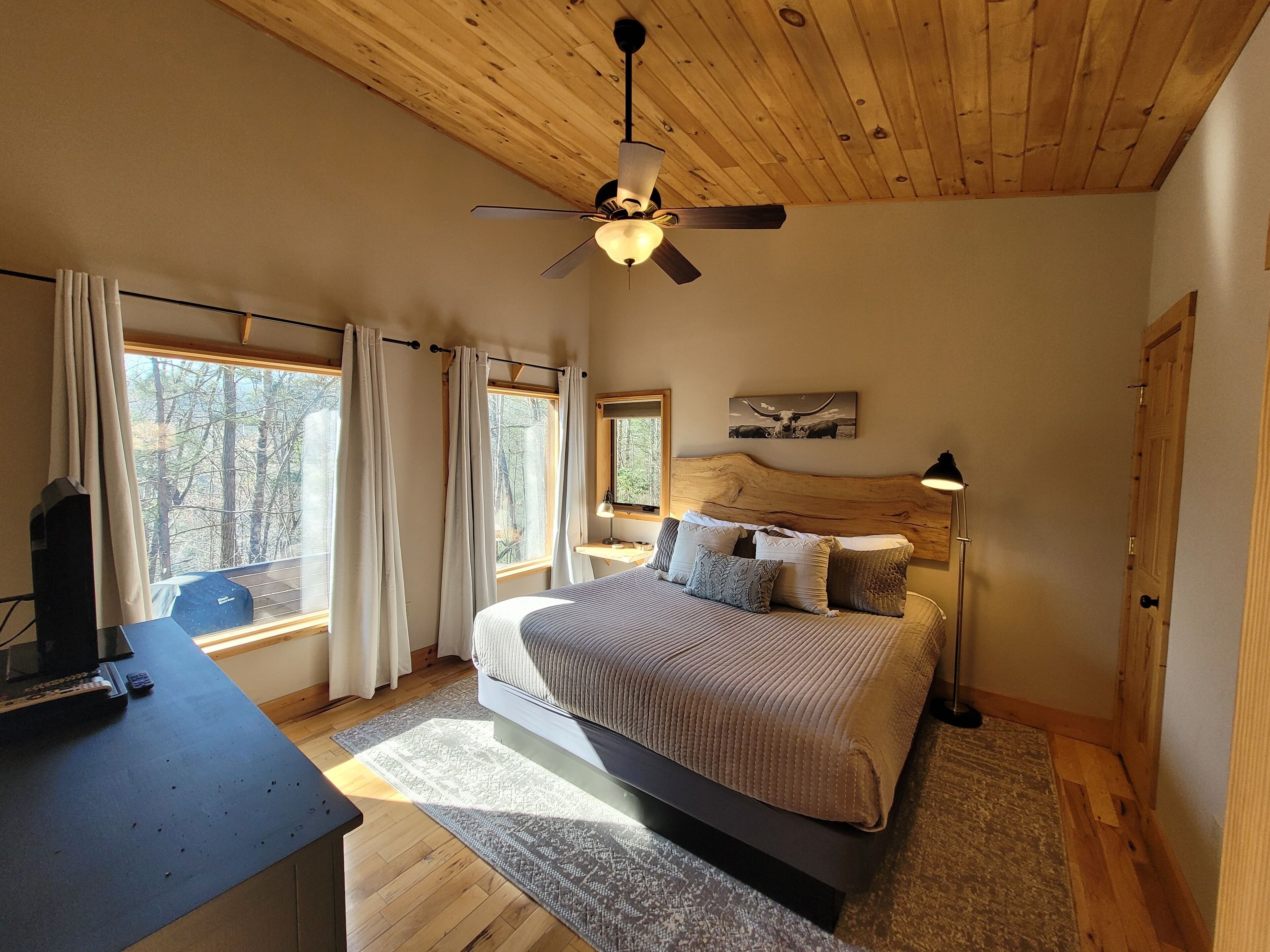 A bedroom with a bed, a desk, a ceiling fan, and windows with curtains.