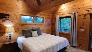 Dormitorio de cabaña de madera con una cama, una mesita de noche, un ventilador de techo y ventanas.