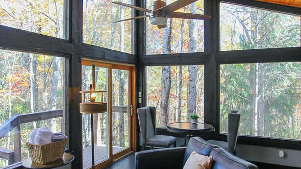 A spacious room with a large window, a ceiling fan, a black leather sofa, and a small table with a basket. - Woodland Loft 1 - Modern-Industrial Mountain Getaway for Two (Bryson City)