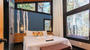 A bedroom with a large bed, a wooden bench, and a view of trees through the windows. - Woodland Loft 1 - Modern-Industrial Mountain Getaway for Two (Bryson City)