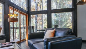 A modern living room with a black leather sofa, a wooden coffee table, and a large window offering a view of a forest. - Woodland Loft 1 - Modern-Industrial Mountain Getaway for Two (Bryson City)