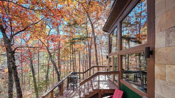 Eine Holzterrasse mit Geländer, zwei rote Adirondack-Stühle und Blick auf einen Wald im Herbstlaub.