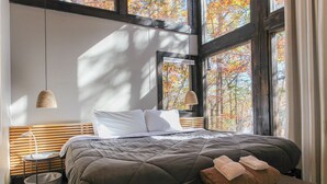 Un dormitorio moderno con una cama grande, una mesita de noche, una lámpara y vistas a árboles otoñales a través de amplias ventanas.