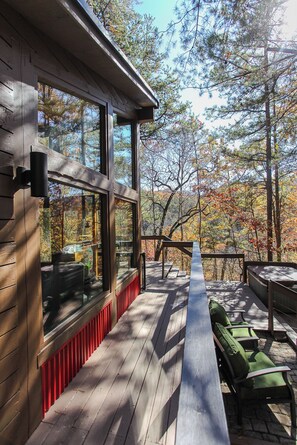 Une terrasse en bois avec un banc et des chaises, de grandes fenêtres et une vue sur les arbres.