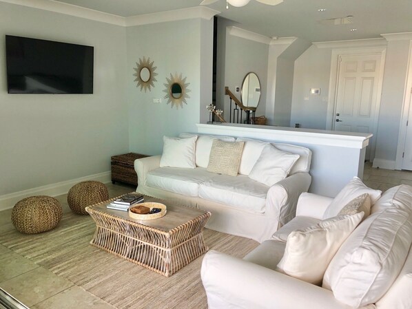 A living room with a white sofa, a wicker coffee table, and a flat-screen TV mounted on the wall.