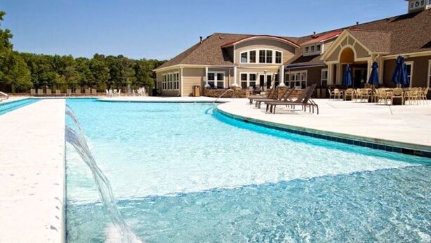 A large swimming pool with a curved edge, surrounded by lounge chairs and umbrellas, and a building with a curved roof and large windows in the background.