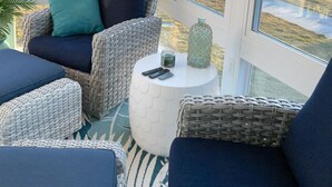 A sunroom with wicker furniture, a small table with a candle and remote controls, and large windows offering a view of a suburban street.