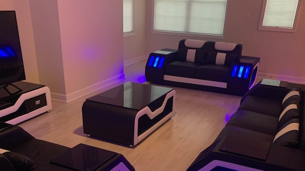 A modern living room with a glass coffee table, black sofas with blue LED lighting, and a flat-screen TV on a black stand.