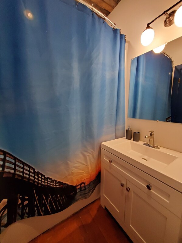 A bathroom with a white vanity, a mirror with lights, a blue shower curtain, and a wooden floor.