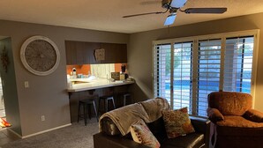 Una sala de estar con un sofá de cuero marrón, un sillón estampado con motivos florales y una zona de cocina con un taburete de bar y un reloj en la pared.