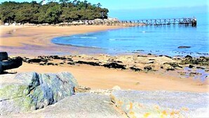 Una playa con un muelle de madera que se adentra en aguas tranquilas, rodeada de rocas y árboles.