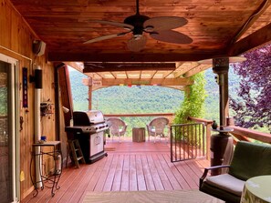 Una terraza de madera con una parrilla, sillas y vistas a una montaña boscosa.