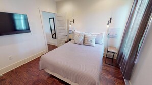 A bedroom with a large bed, a flat-screen TV mounted on the wall, a bedside table with a lamp, and a door leading to another room. - The Seahorse 30A! Golf Cart Included! Sleeps 10! (Santa Rosa Beach)