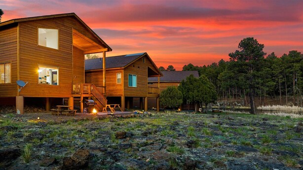 Una cabaña de madera de dos pisos con terraza y chimenea, rodeada por un paisaje natural.