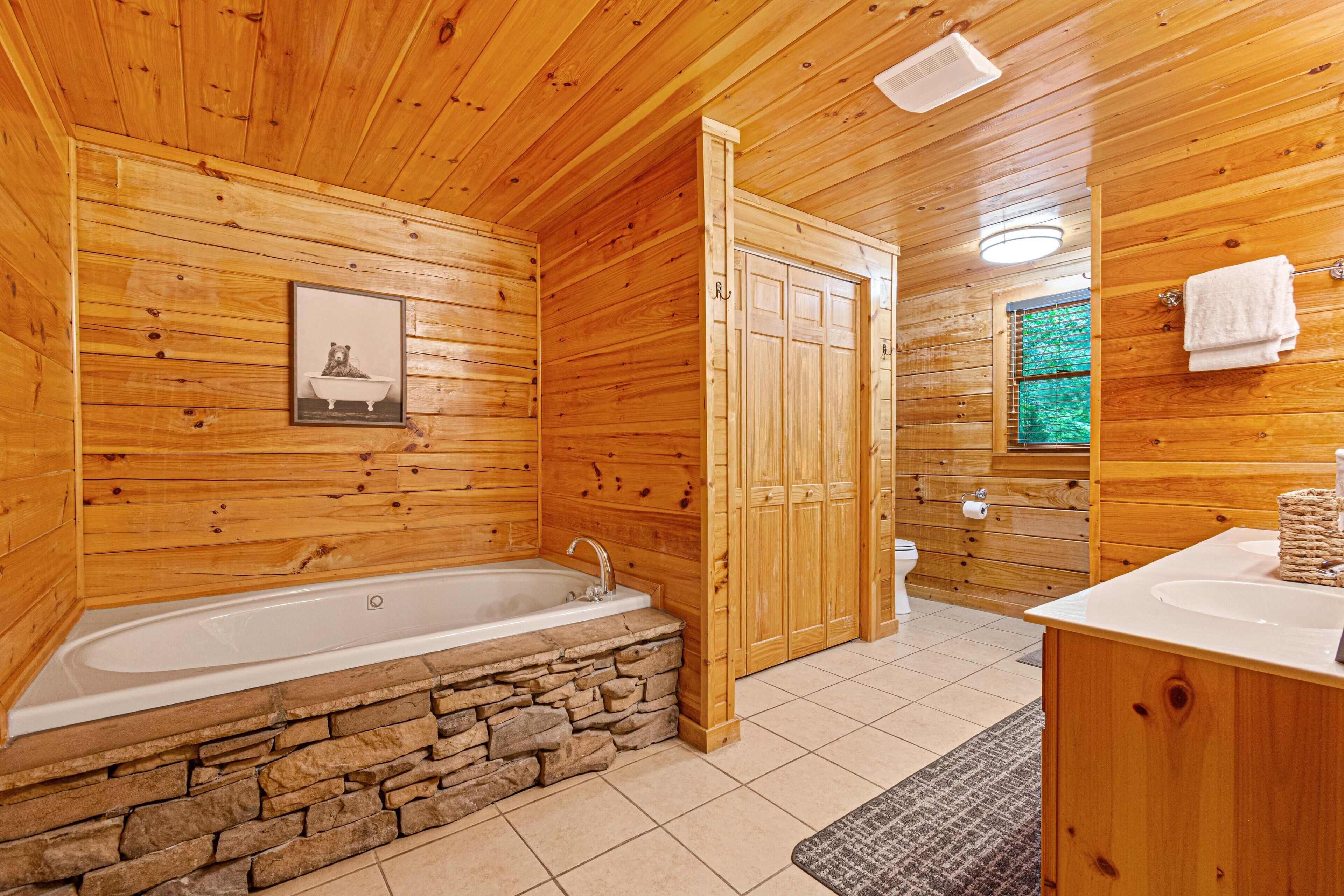 A wooden cabin bathroom with a stone-lined bathtub, wooden cabinets, and a window with a view of greenery.
