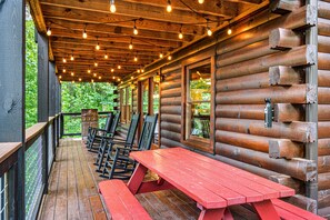 Una terraza de madera con una mesa y sillas de picnic rojas, rodeada de paredes de cabaña de troncos y grandes ventanales.