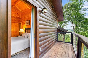 Un dormitorio de cabaña de troncos con una cama, una mesita de noche, una lámpara y una ventana con vista a un área boscosa.