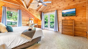 Un dormitorio con cama, ventilador de techo, televisor y vistas al exterior a través de amplias ventanas.