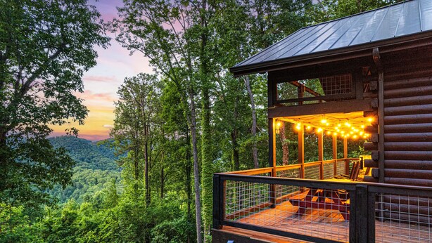 Una cabaña de madera con terraza y luces cálidas, rodeada de un bosque frondoso y vistas a las montañas.