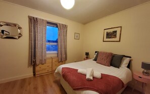 A bedroom with a bed, a window with curtains, a bedside table, a lamp, and a framed picture on the wall.