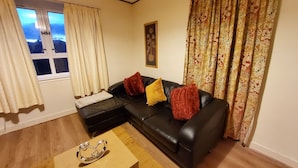 Sala de estar com um sofá de couro preto, duas almofadas vermelhas, uma almofada amarela e uma mesa de centro de madeira. Há uma janela com cortinas brancas, um quadro na parede e cortinas estampadas à direita.