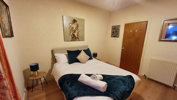 Cama bem arrumada com manta azul e toalha enrolada, mesa de cabeceira com abajur e quadro na parede.