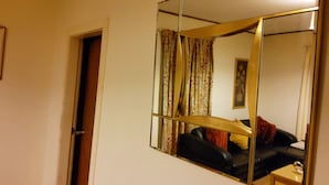 Quarto com um espelho grande refletindo um sofá de couro preto, uma mesa pequena e uma porta.
