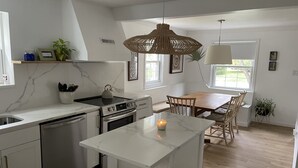 Uma cozinha moderna com bancadas de mármore branco, fogão embutido e um espaço de refeições com cadeiras e mesa de madeira.