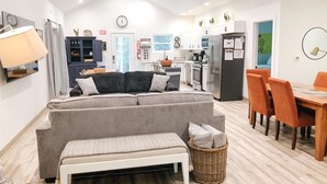 A modern apartment with a living area featuring a grey sofa, a dining table with orange chairs, a kitchen with white cabinets, and a visible clock on the wall. - Eagle's Nest Guest House II - Less than 2 Miles to Sand Valley - 16 beds! (Nekoosa)