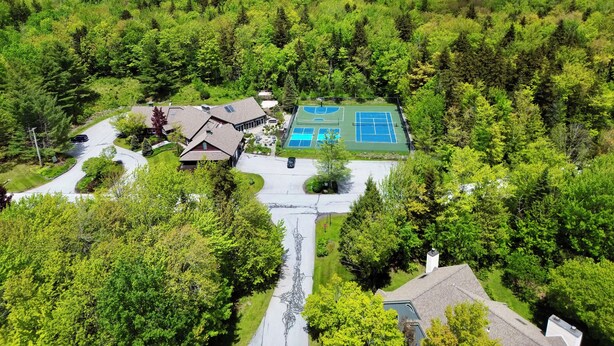 Una zona residencial con una casa, un camino de entrada y una cancha de tenis, todo rodeado de un bosque denso.
