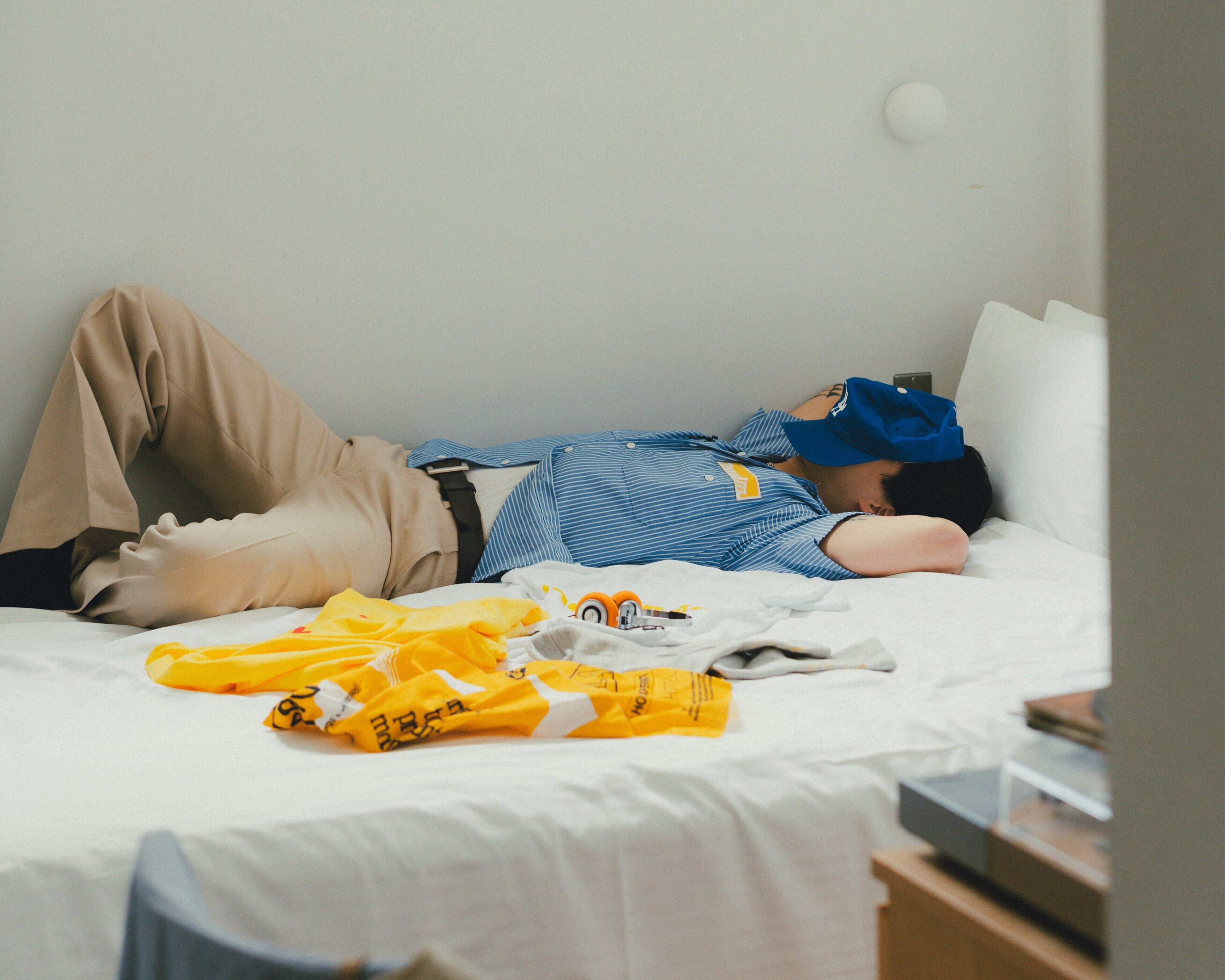 A person lying on a bed with a blue and white striped shirt, beige pants, and a blue cap.