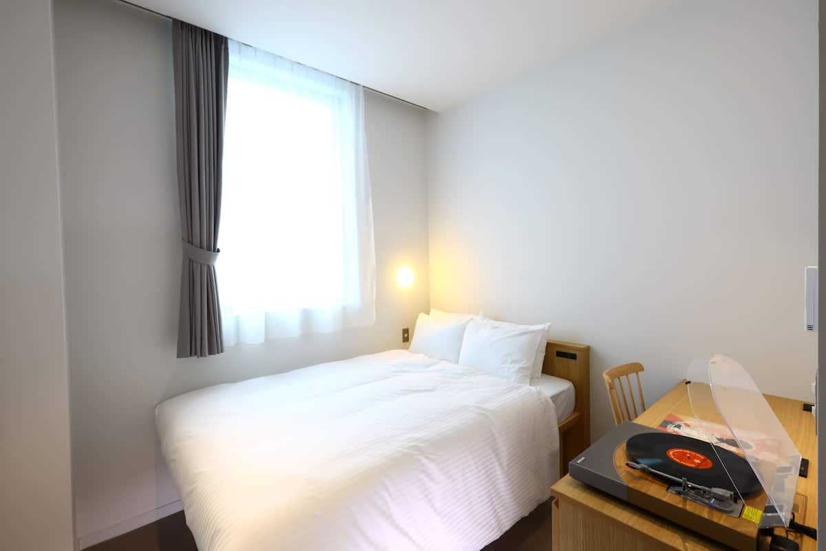 A hotel room with a bed, a desk with a record player, a chair, and a window with curtains.