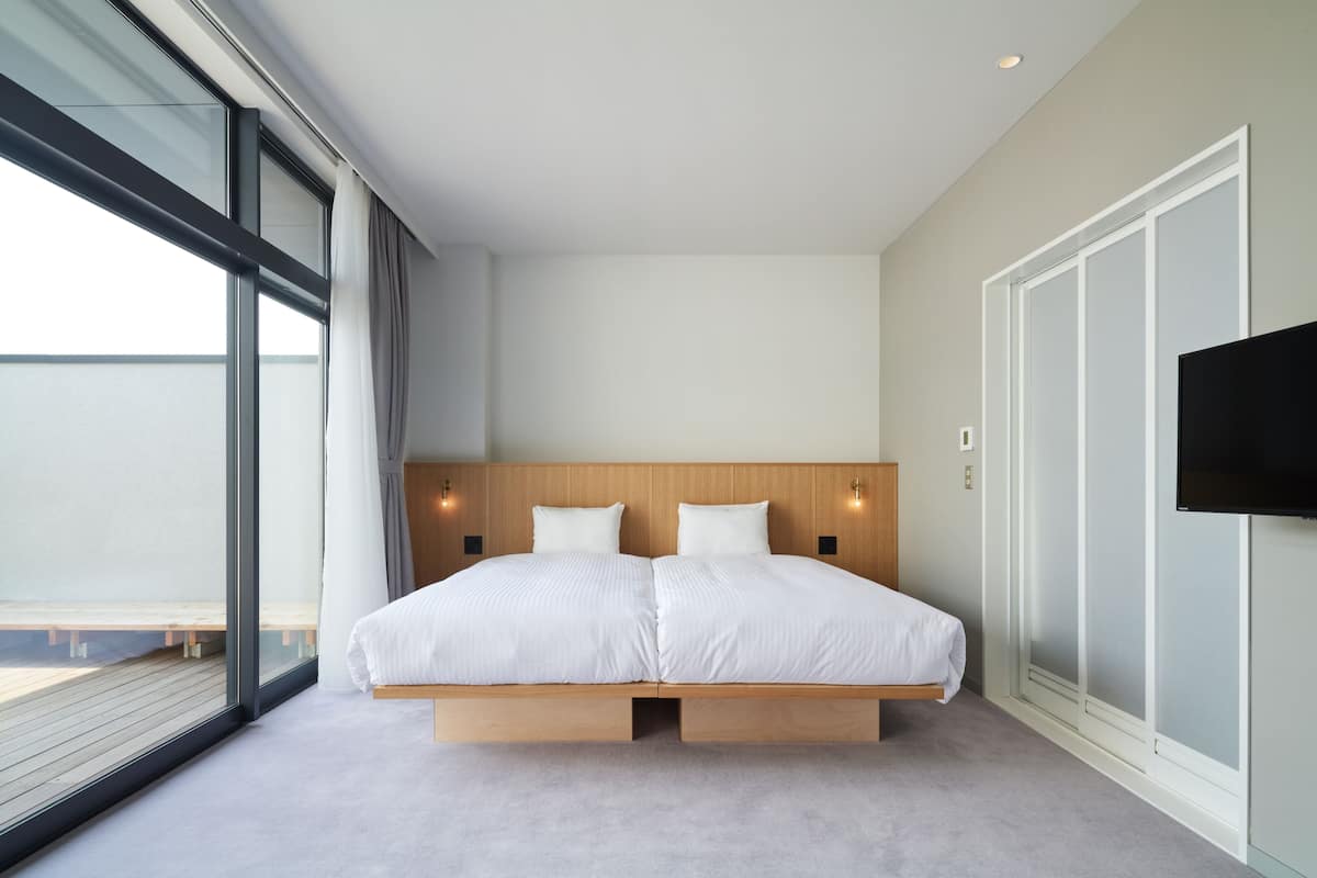 A modern bedroom with a large bed, a TV mounted on the wall, and a view of the outdoors through large windows.
