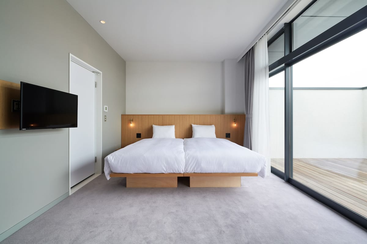A modern bedroom with a large bed, a flat-screen TV mounted on the wall, and a large window offering a view of the outdoors.