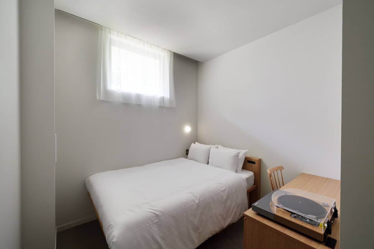 A small, neatly arranged bedroom with a single bed, a wooden nightstand, a table with a turntable, and a window with sheer curtains.