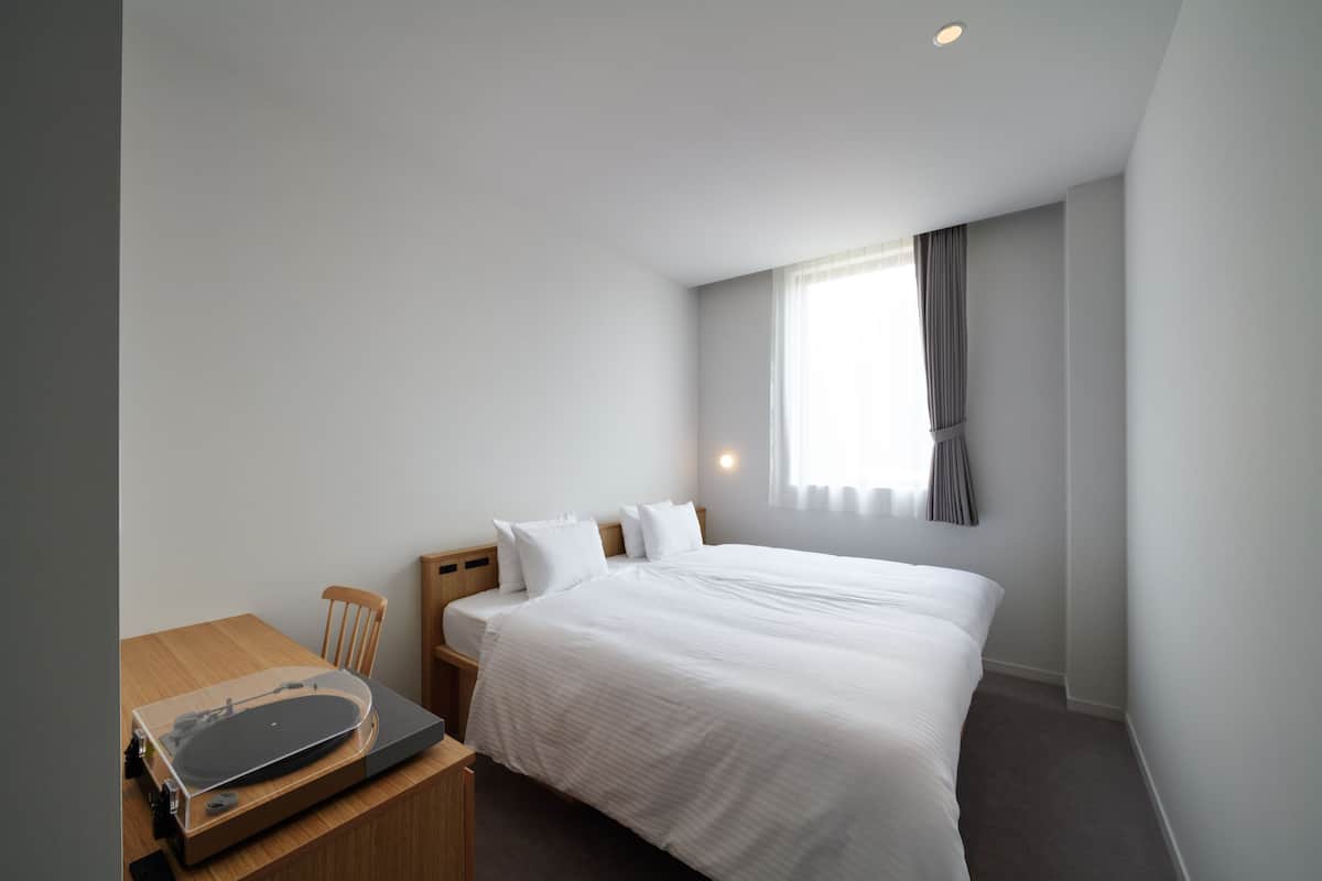 A hotel room with a bed, a desk with a turntable, a chair, and a window with curtains.
