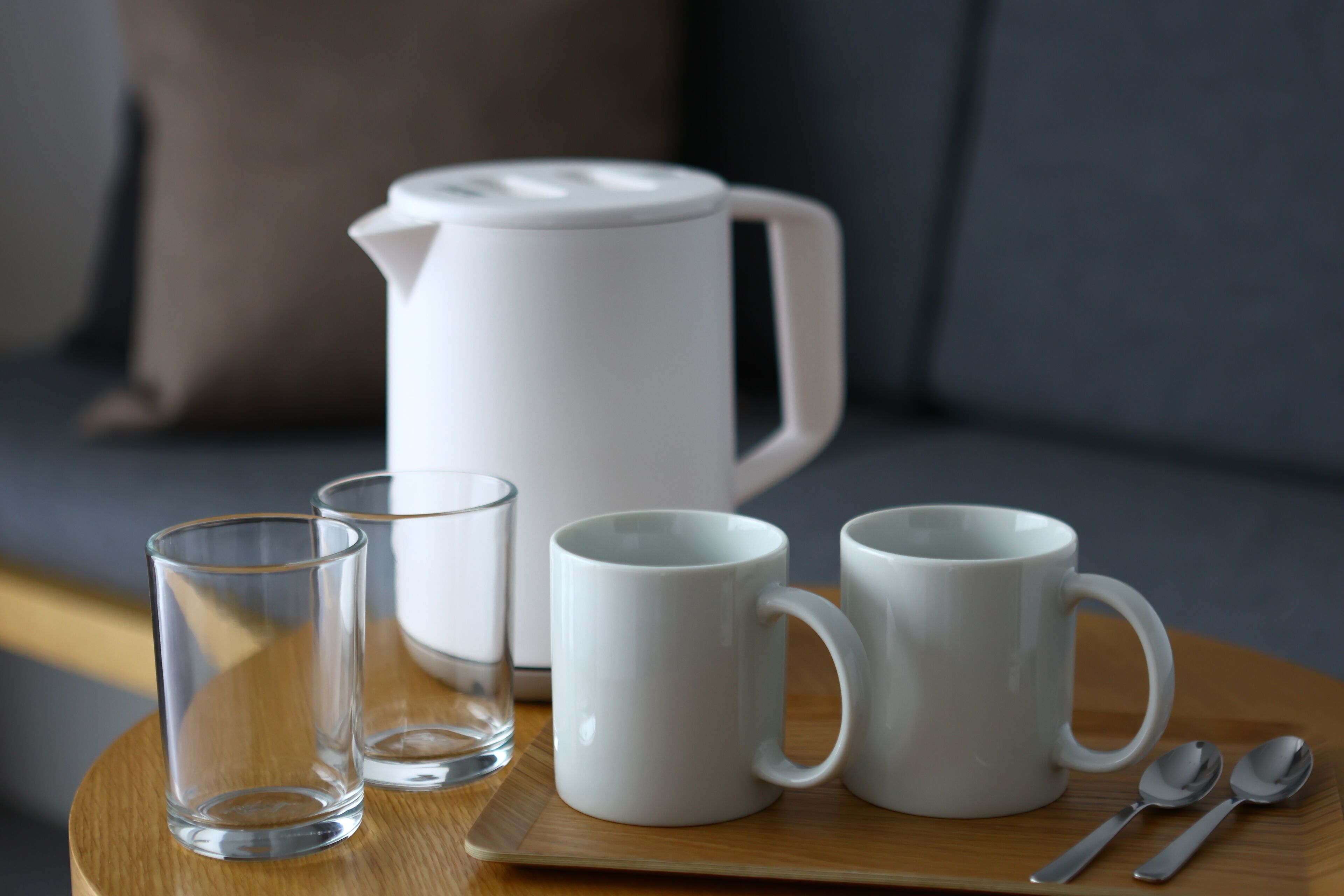 A tray with two clear glasses, two white mugs, a white teapot, and two spoons on a wooden surface.