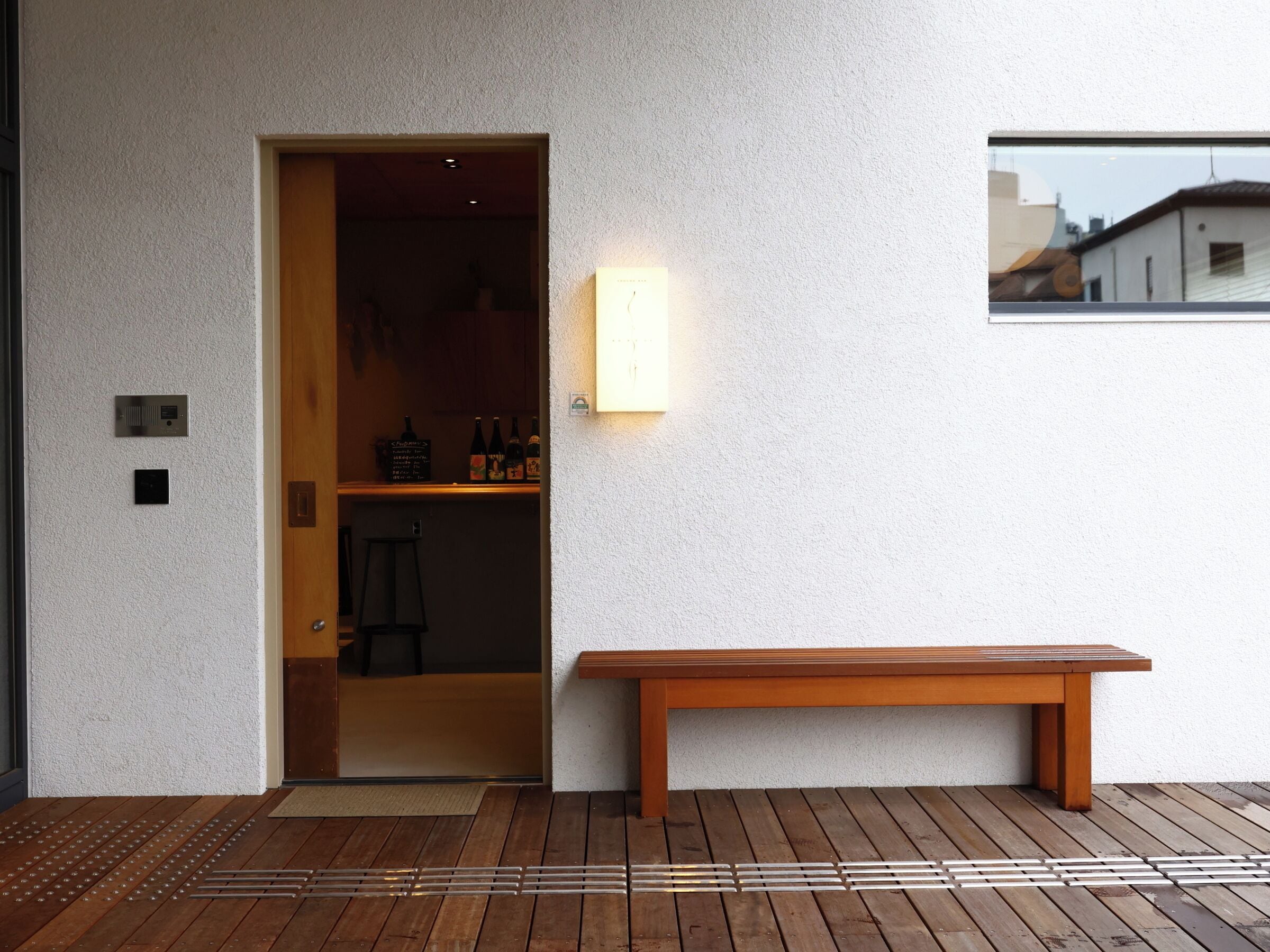 A wooden bench outside a building with a white textured wall, a door leading to an interior space, and a small window reflecting a building.