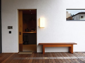A wooden bench outside a building with a white textured wall, a door leading to an interior space, and a small window reflecting a building. - Mustard Hotel Shimokitazawa (Tokyo)