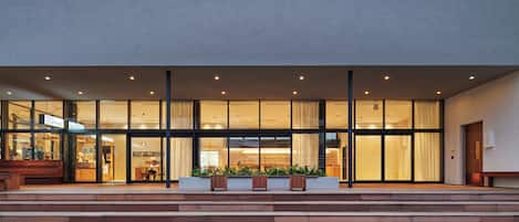 A modern building with a glass facade, wooden steps leading to the entrance, and outdoor seating.