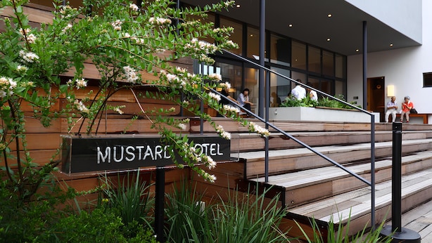 A modern hotel entrance with a wooden staircase, a sign reading "MUSTARD HOTEL," and a view of the interior through large windows.