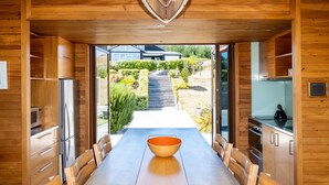 A modern kitchen with wooden cabinetry, a central island with a bowl, and a view of a house and garden through an open door. - Bach or holiday home, 1 Bedrooms, 2 Bathrooms, (Sleeps 2) (Waimarama)